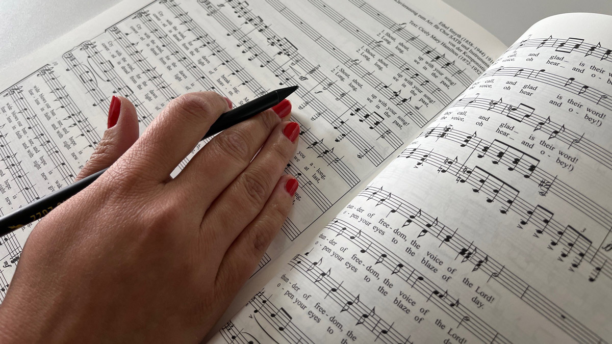 Ein Blatt mit Noten eines Liedes, darüber die Hand einer Frau mit einem Stift. Das Lied ist von einer Frau komponiert und trägt den englischen Titel The March of the Women, der Marsch der Frauen.