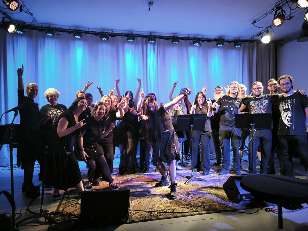 Mehrere Menschen stehen auf einer blau angeleuchteten Bühne. Viele tragen schwarze Kleidung und zeigen den Heavy Metal Gruß mit ihren Händen.