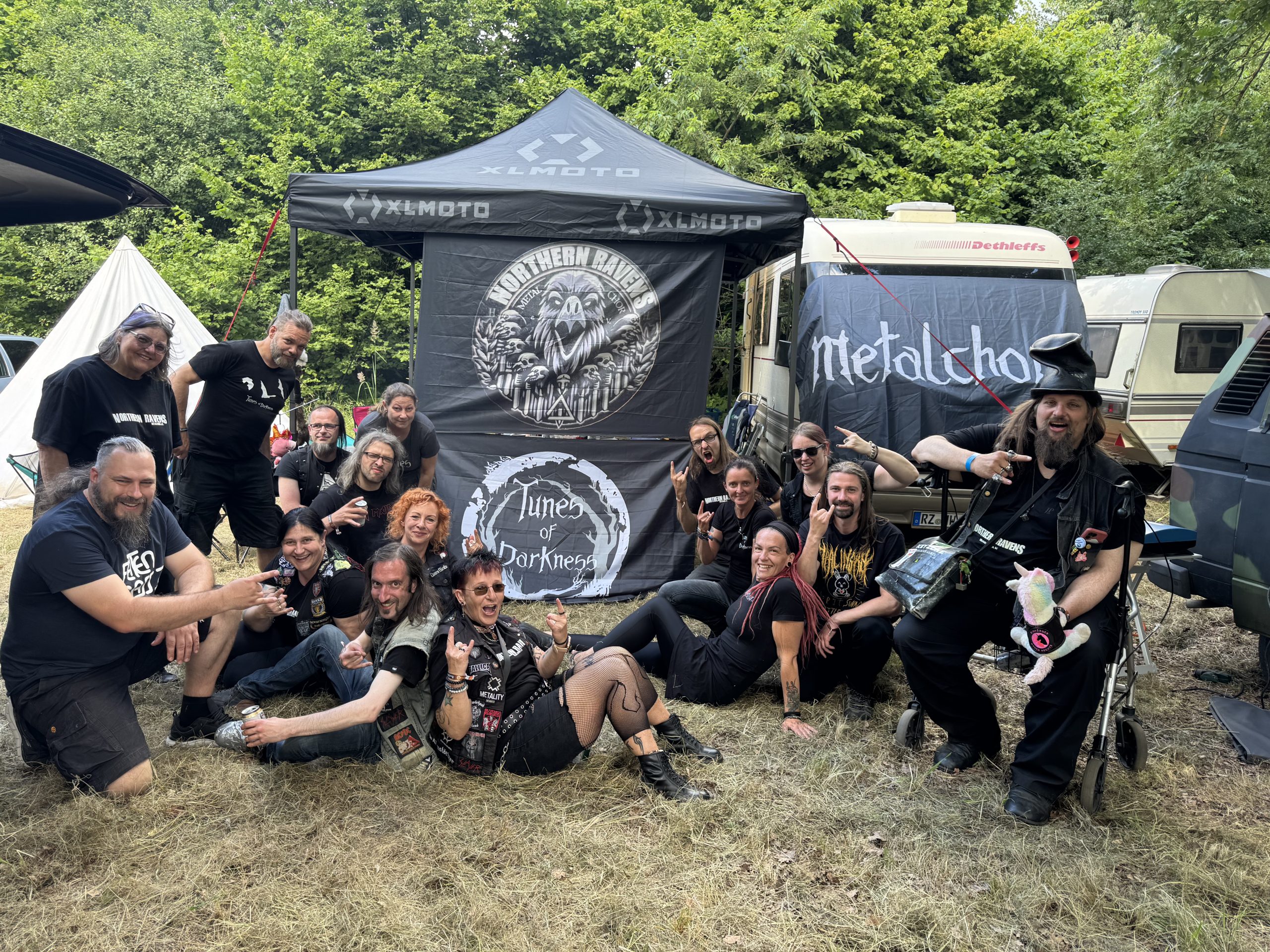 Etwa 15 Menschen sitzen, stehen und liegen vor einem Zelt, das mit den Logos der Heavy-Metal-Chöre geschmückt ist. Die Menschen schauen lächelnd in die Kamera, einige machen Grimassen. Sie tragen schwarze Kleidung und teilweise Heavy Metal T-Shirts.