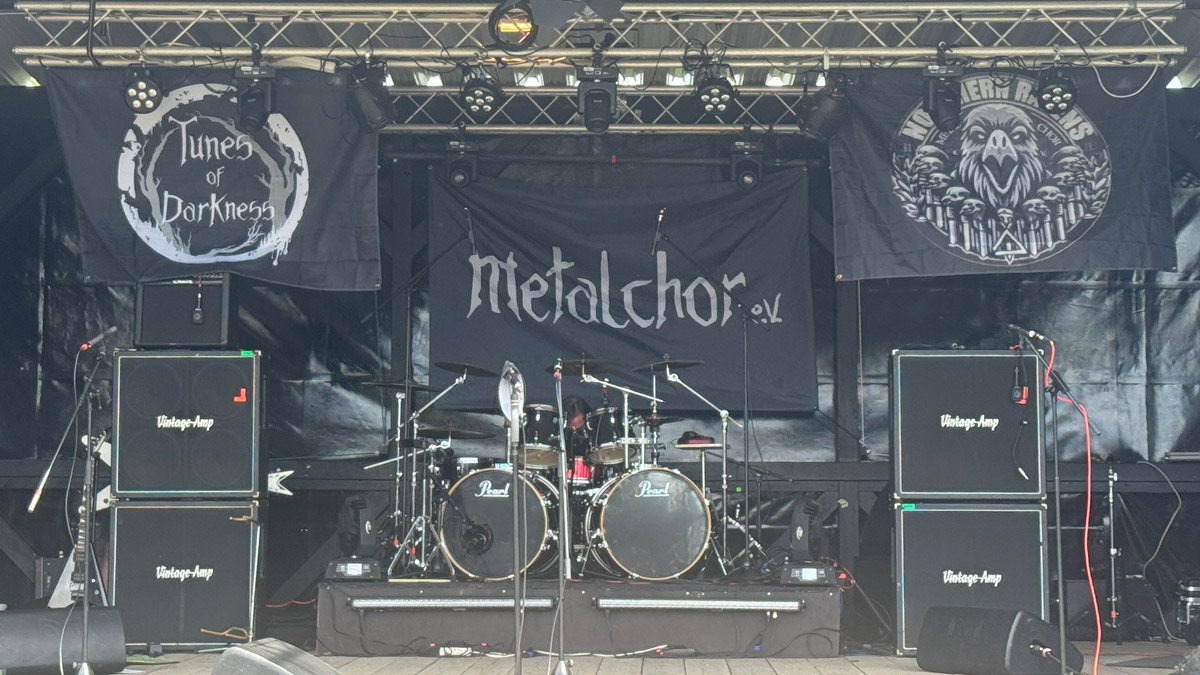 Eine leere Open-Air-Bühne mit Schlagzeug, mehreren Verstärkern und Mikrofonständern. Über der Bühne hängen Banner mit den Schriftzügen „Tunes of Darkness“, „Metalchor e.V.“ und „Northern Ravens“. Die Bühne ist bereit für einen Auftritt.