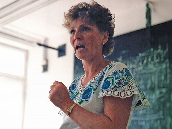 Eine Frau mit hellbrauen, lockigen Haaren steht vor einer Tafel und spricht