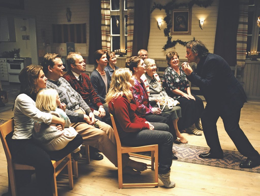 Eine Gruppe von Menschen sitzt in einem gemütlich eingerichteten Raum auf Stühlen in einer Reihe. Ein Mann steht vor ihnen und spricht gestikulierend zu der Gruppe. Im Hintergrund sind eine Küche, ein Fenster mit Vorhängen, Kerzen und ein Bild an der Wand zu sehen. Die Atmosphäre wirkt warm und familiär.