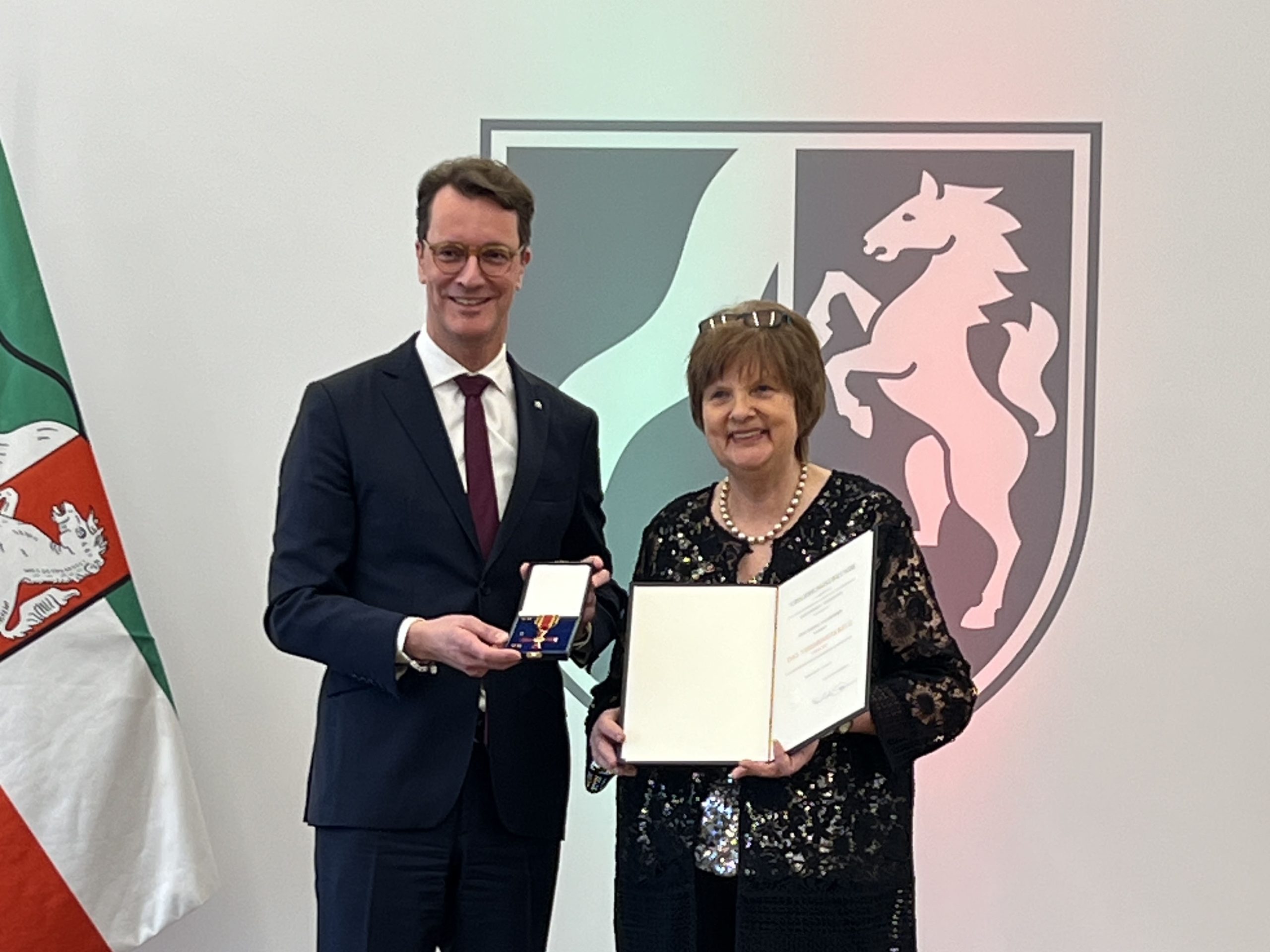 Eine Frau, die lächelnd mit einer Urkunde in der Hand vor dem Wappen Niedersachsens steht, erhält von einem großen Mann neben ihr einen Orden.