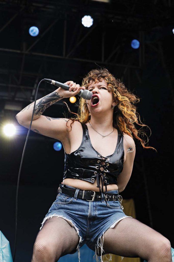 Eine Frau mit lockigen blond-roten Haaren steht mit Mikro in der Hand auf der Bühne. Sie schreit kraftvoll in das Mikrofon. Sie trägt eine kurze Lederweste, kurze Jeans und Netzstrümpfe.