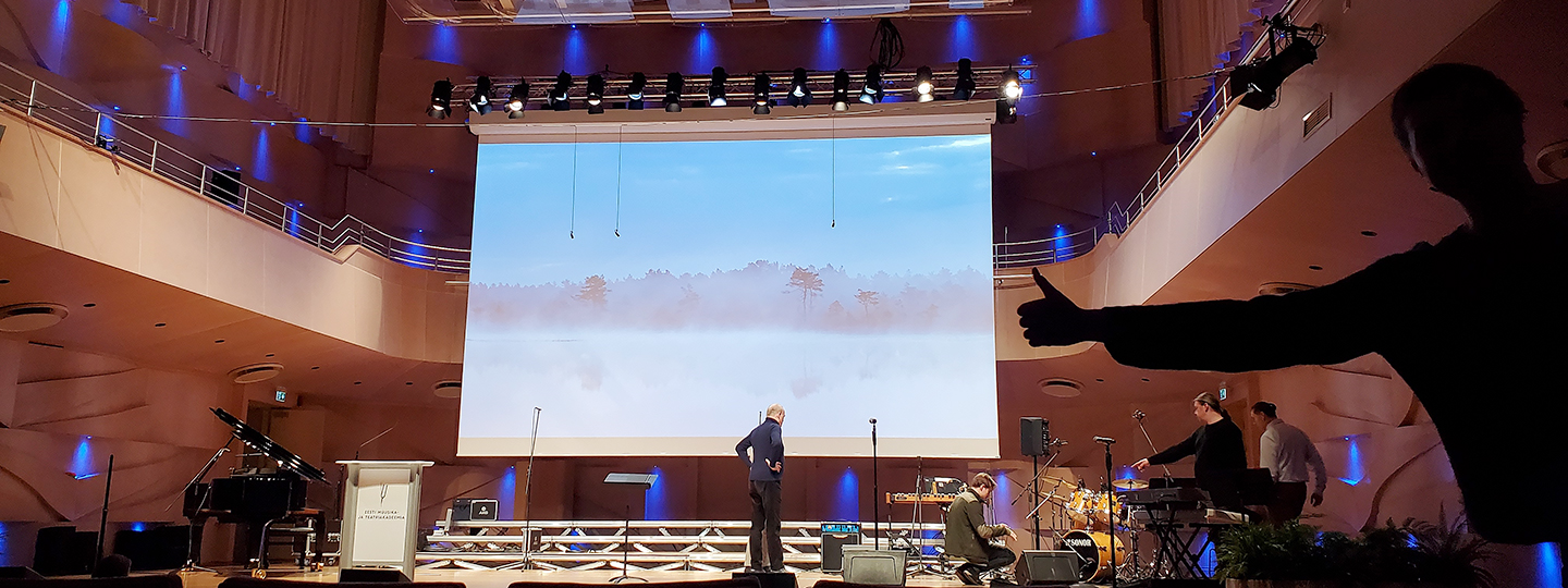 Eine Leinwand, angebracht auf der Bühne eines Konzertsaals, zeigt eine Landschaft mit Bäumen im Nebel.