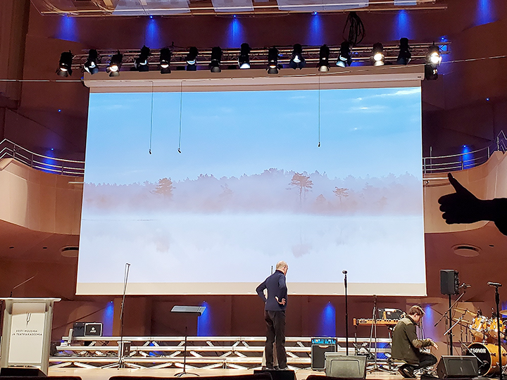 Eine Leinwand, angebracht auf der Bühne eines Konzertsaals, zeigt eine Landschaft mit Bäumen im Nebel.