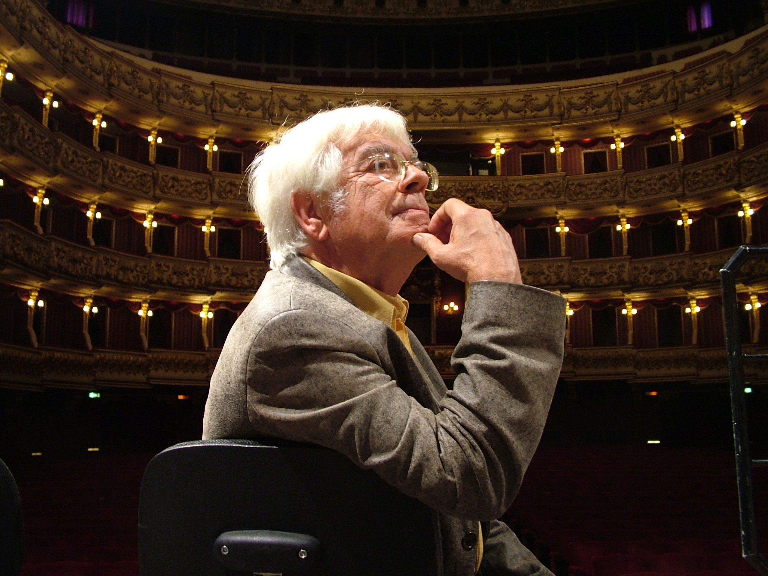 Helmuth Rilling sitzt im Profil auf einem Stuhl, im Hintergrund ein festlicher Theaterraum