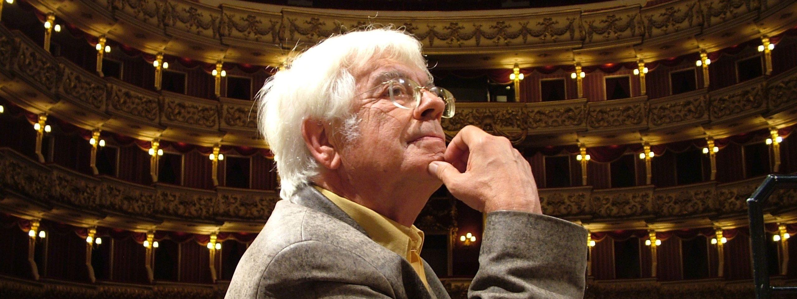 Helmuth Rilling sitzt im Profil auf einem Stuhl, im Hintergrund ein festlicher Theaterraum