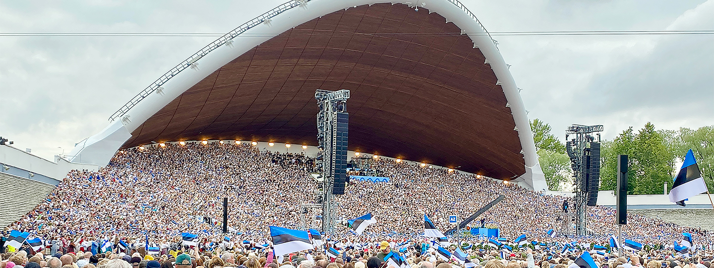 Tausende von Menschen stehen auf und vor einer sehr großen überdachten Open-Air-Bühne.