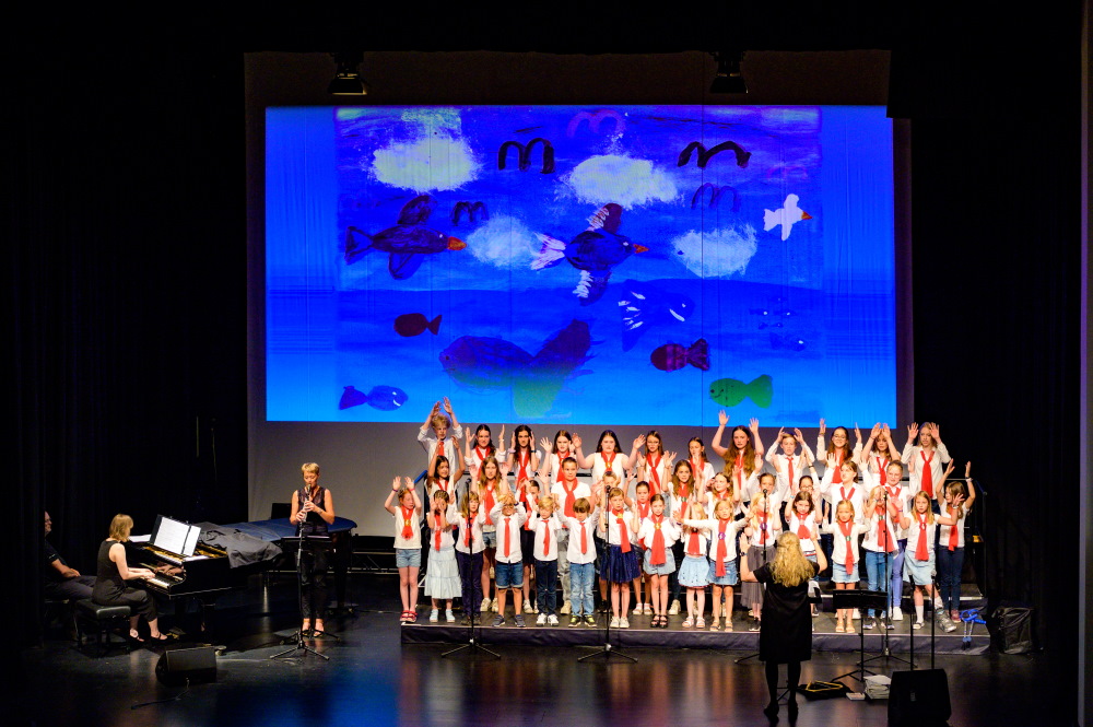 Auf einer Bühne singt und agiert ein Kinderchor, im Hintergrund zeigt eine Leinwand Fische und Vögel