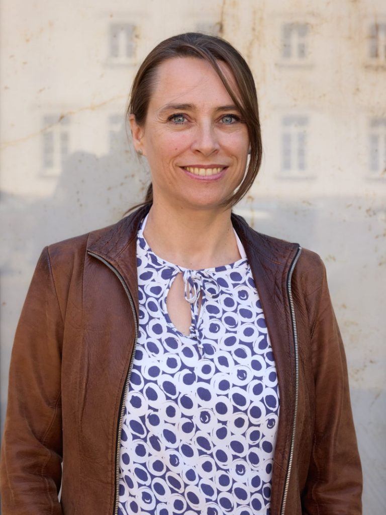 Ein Portrait der Studiengangsbeauftragten Professorin Anne Steinbach in brauner Lederjacke vor reflektierendem Hintergrund.