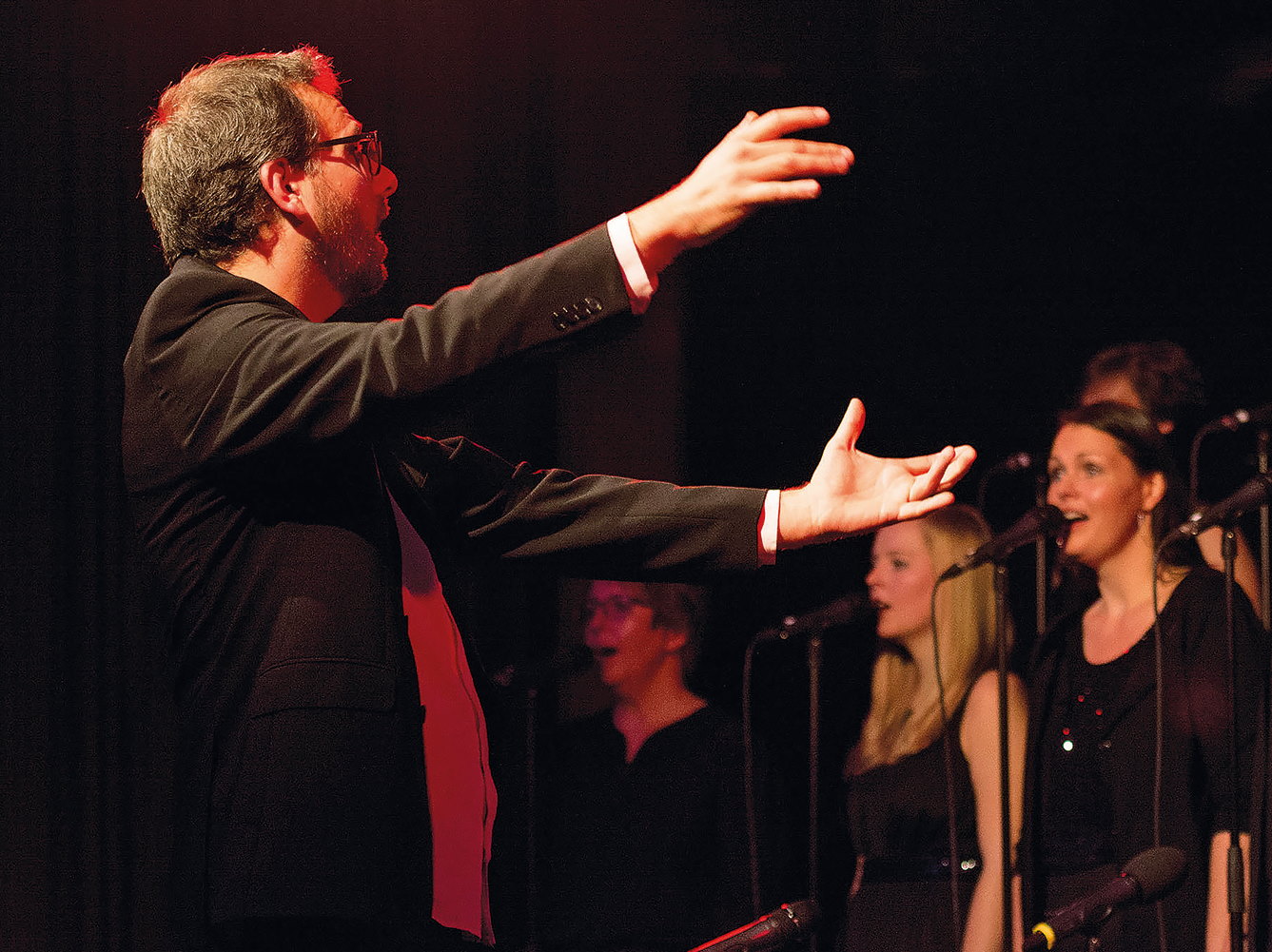 Ein Chorleiter mit weit ausgestreckten Armen vor einigen Frauen aus dem Chor