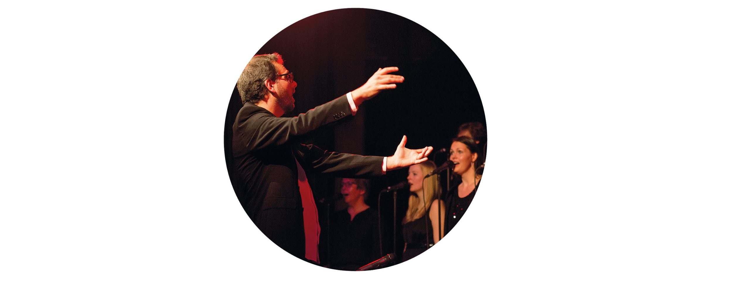 Ein Chorleiter mit weit ausgestreckten Armen vor einigen Frauen aus dem Chor