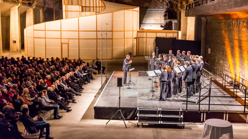 Ein kleiner Chor steht in einer großen Halle rechts auf einer Bühne, links sitzt das Publikum.