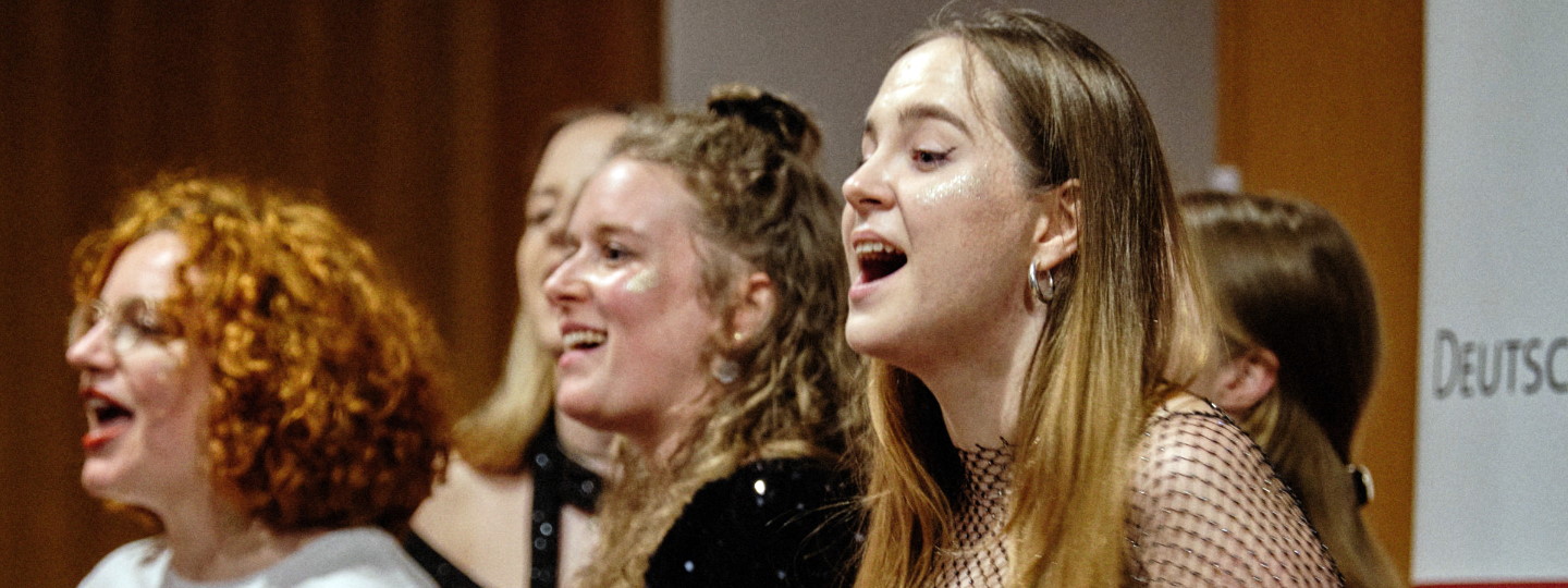 Chorsängerinnen in Aktion, im Hintergrund Wand und ein roter Vorhang.