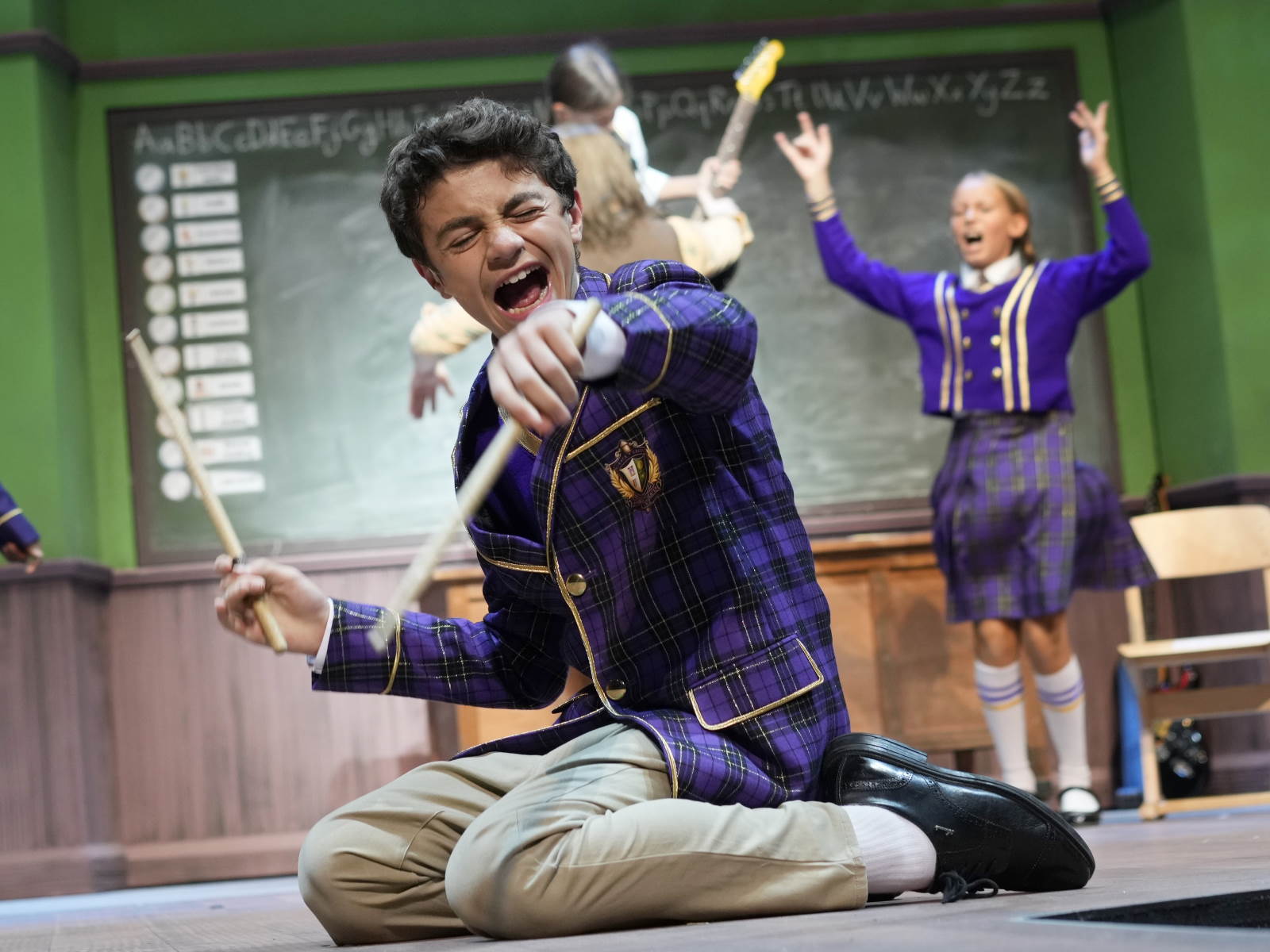 Ein Junge in einer wohl fiktiven Schuluniform kniet in den Kulissen eines Klassenzimmers und singt so leidenschaftlich, dass er die Augen geschlossen hat. In seinen Händen hat er Drumsticks.