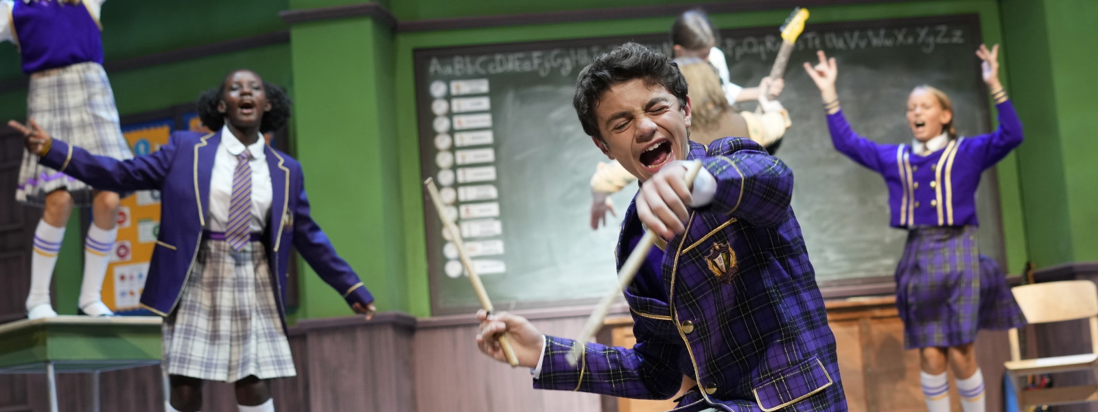 Ein Junge in einer wohl fiktiven Schuluniform kniet in den Kulissen eines Klassenzimmers und singt so leidenschaftlich, dass er die Augen geschlossen hat. In seinen Händen hat er Drumsticks.