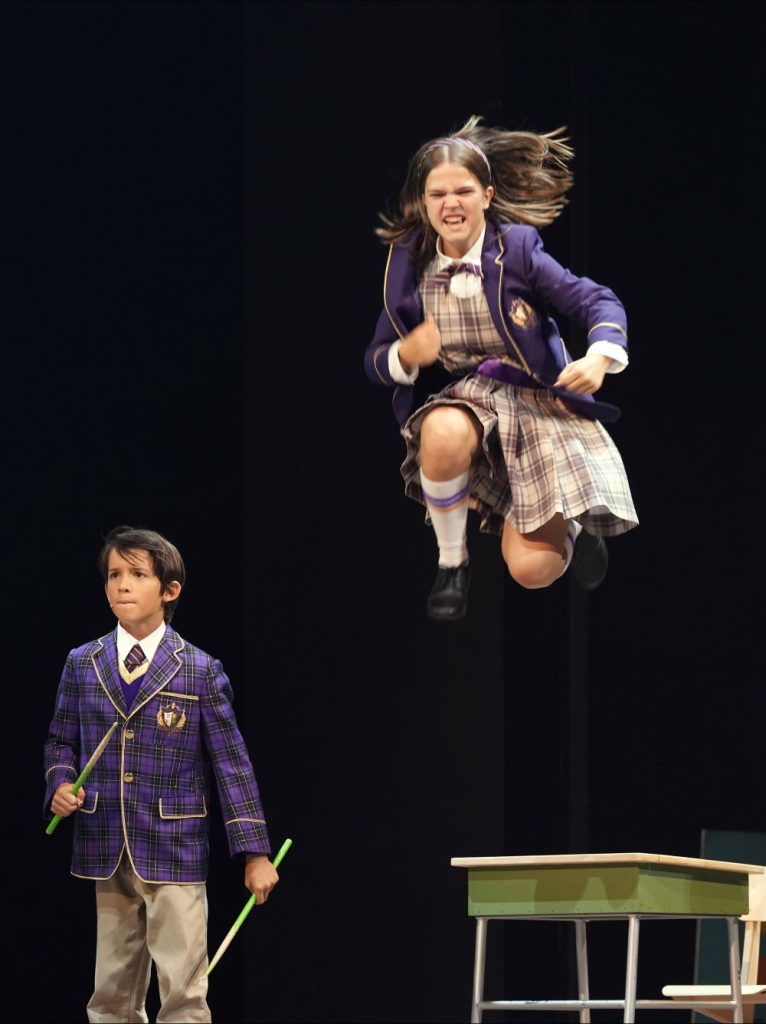 Ein Mädchen in wohl fiktiver Schuluniform springt weit über ein Schulpult und hat dabei das Gesicht energisch angespannt. Ein weiter entfernt stehender Junge hält Drumsticks in den Händen und guckt zaghafter.