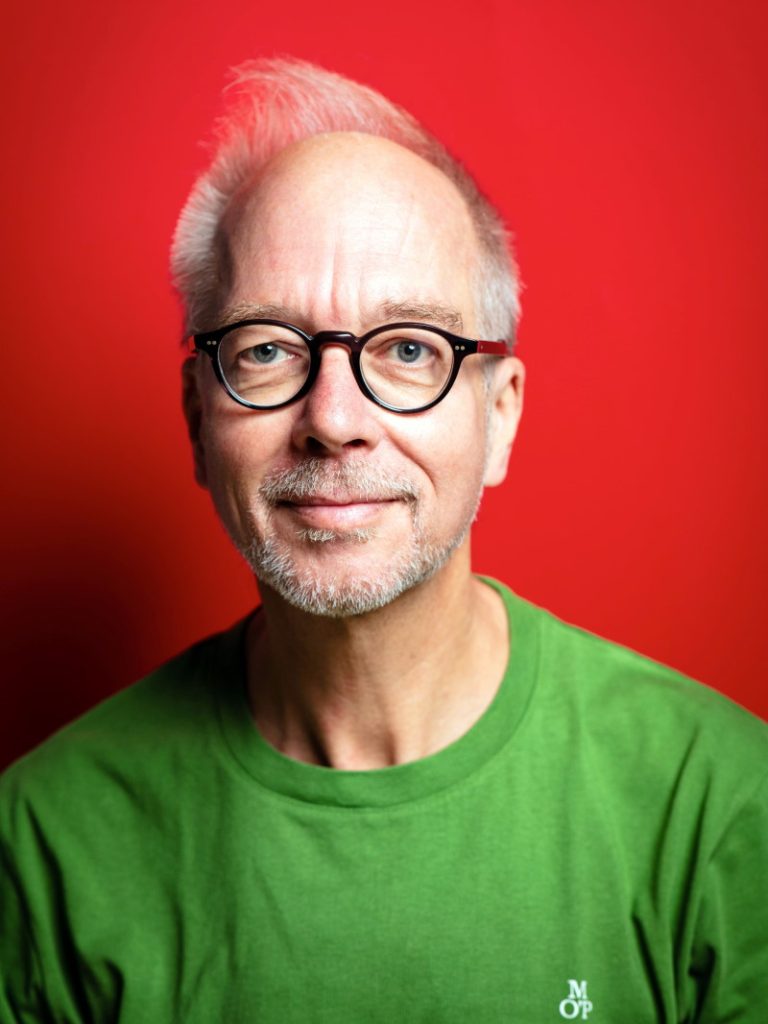 Der Regisseur Matthias Davids in Nahaufnahme vor einer roten Wand. Er trägt ein grünes T-Shirt und blickt, minimal lächelnd, durch die Gläser seines dick schwarz umdandeten Brillengestells.