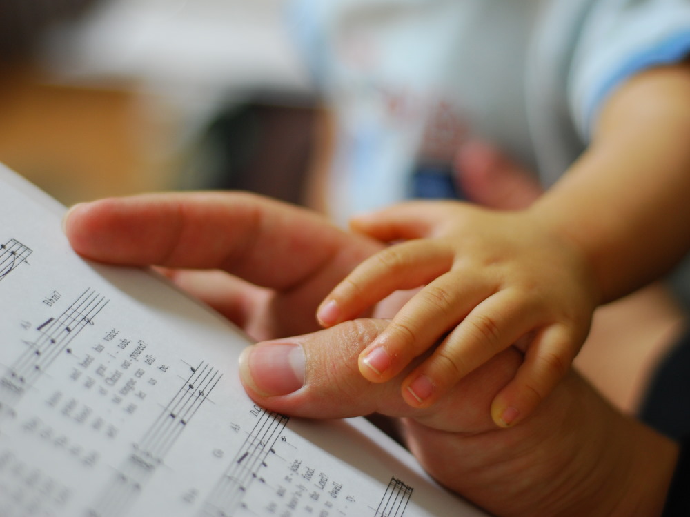 Die Hand einer erwachsenen Person hält ein Notenblatt. Ein kleines Kind hat seine Hand auf die Hand der erwachsenen Person gelegt.