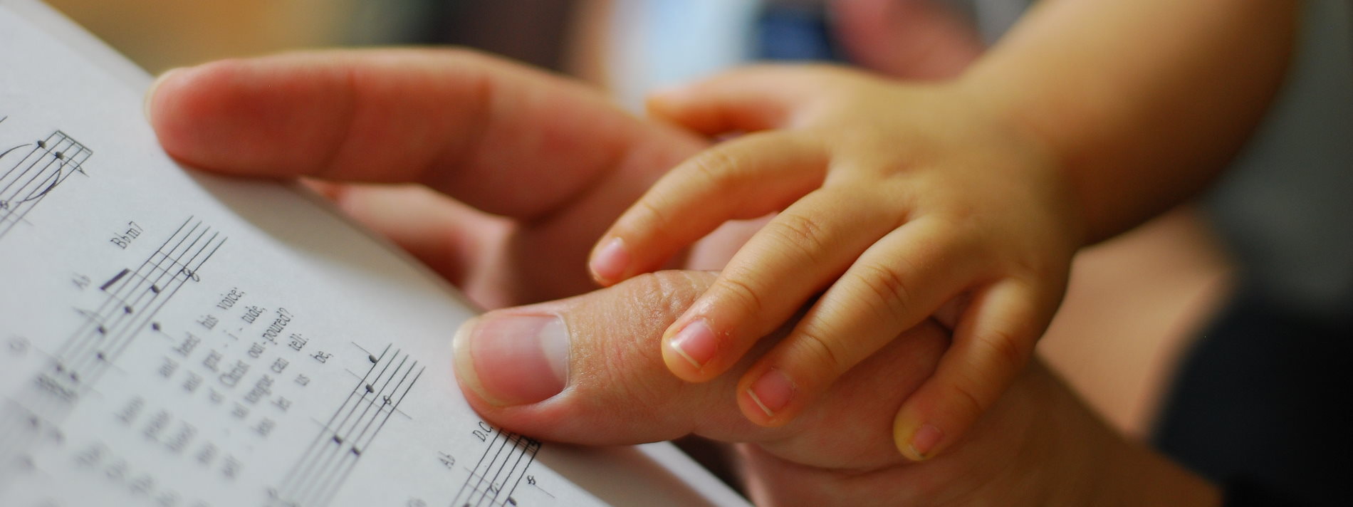 Die Hand einer erwachsenen Person hält ein Notenblatt. Ein kleines Kind hat seine Hand auf die Hand der erwachsenen Person gelegt.