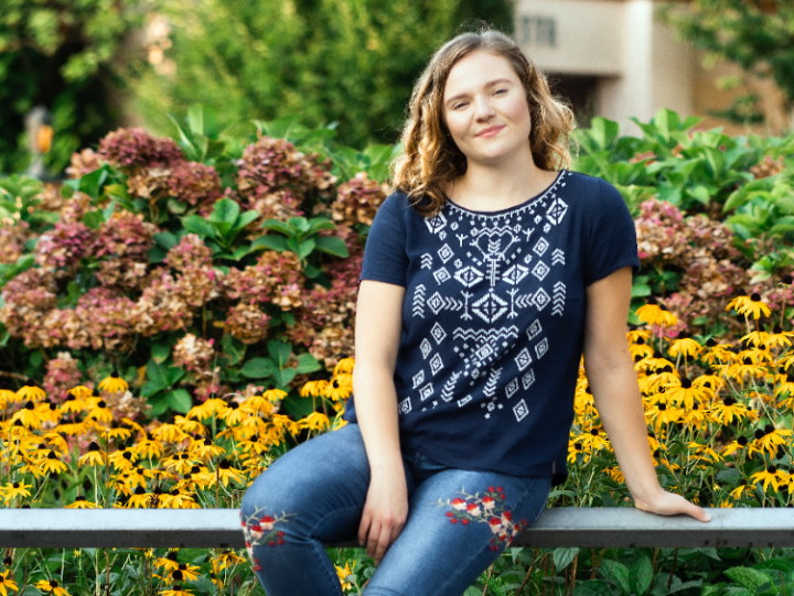 Die Darstellerin Mascha Kamenskikh sitzt auf einer Bank vor einem Beet voller gelb und rot blühender Blumen. Sie hat den linken Arm auf der Bank abgestützt und schaut mit geneigtem Kopf entspannt in die Kamera.