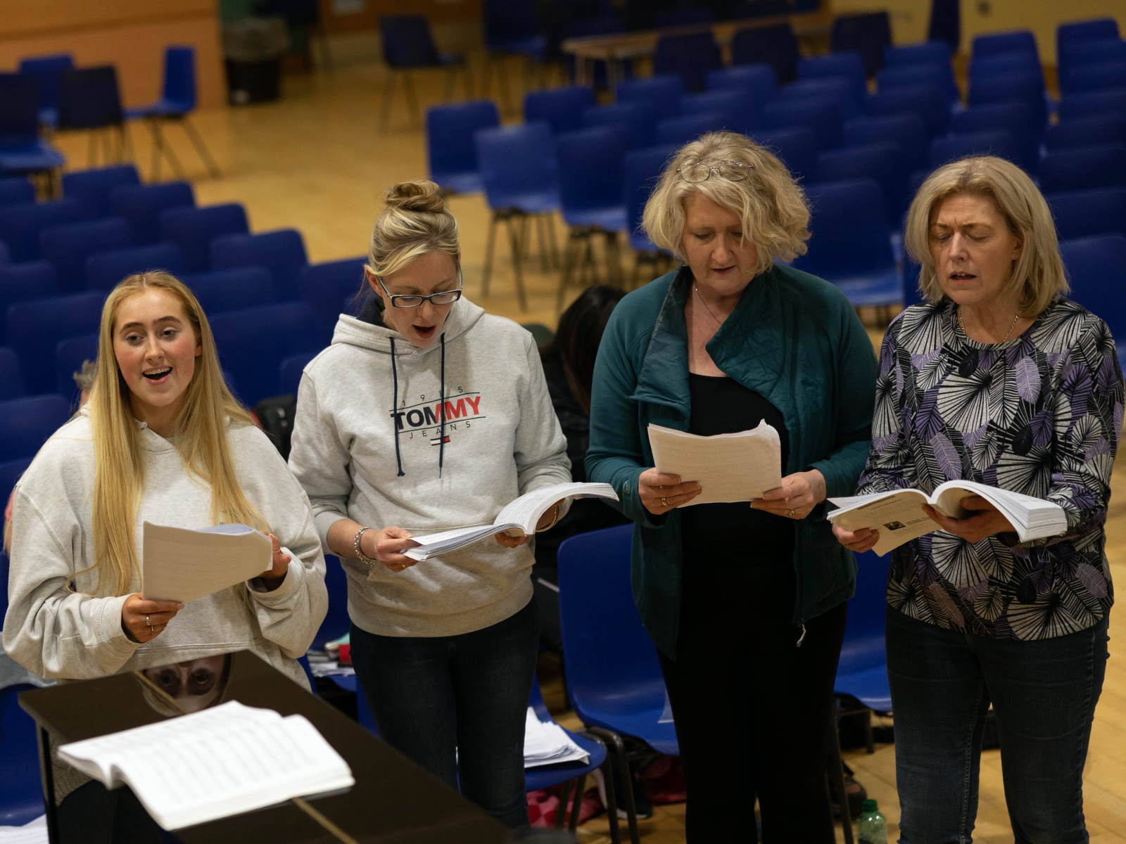 Vier Sängerinnen verschiedenen Alters stehen vor blauen Stuhlreihen auf einer Bühne an einem Klavier und singen. Drei schauen in die Notenausgaben in ihren Händen, die jüngste blickt lächelnd nach vorn.