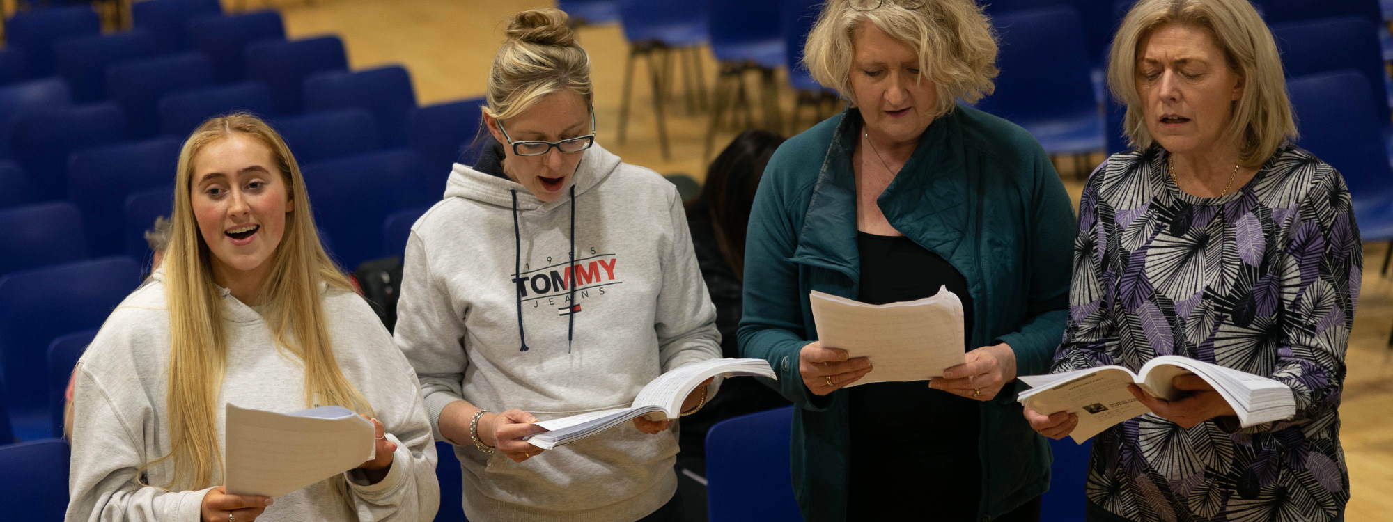 Vier Sängerinnen verschiedenen Alters stehen vor blauen Stuhlreihen auf einer Bühne an einem Klavier und singen. Drei schauen in die Notenausgaben in ihren Händen, die jüngste blickt lächelnd nach vorn.