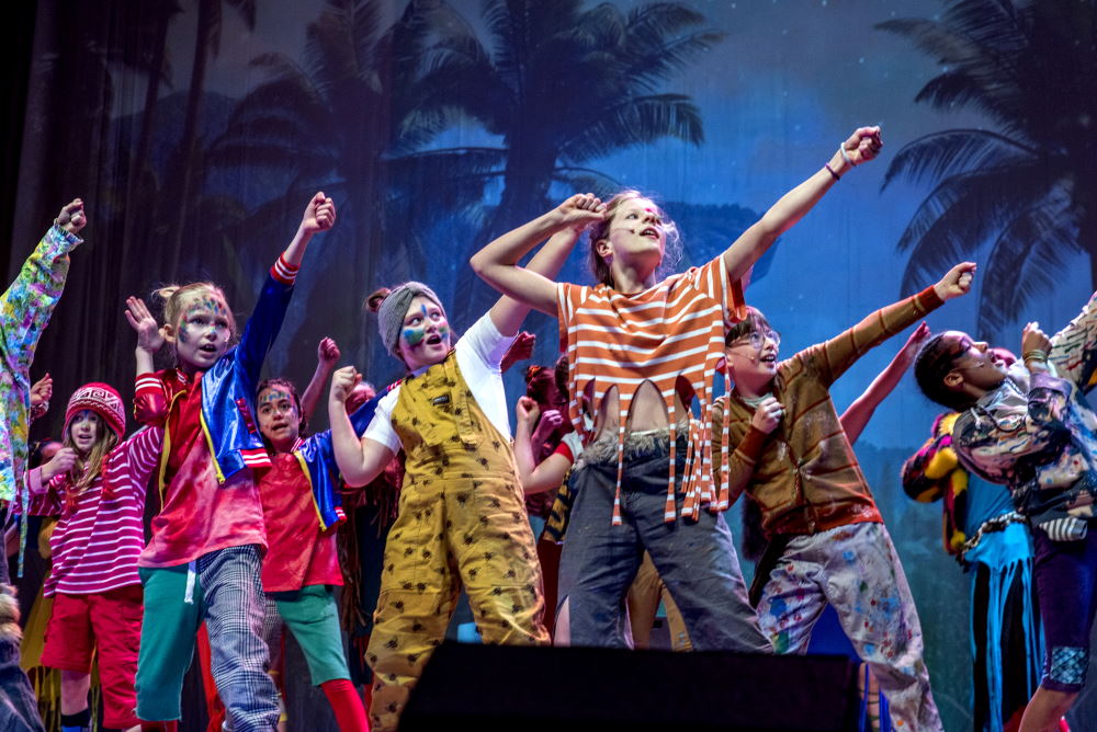 Bunt gekleidete Kinder auf einer Bühne haben die Arme in kraftvoller Pose erhoben, als würden sie Pfeil und Bogen in den Himmel richten.