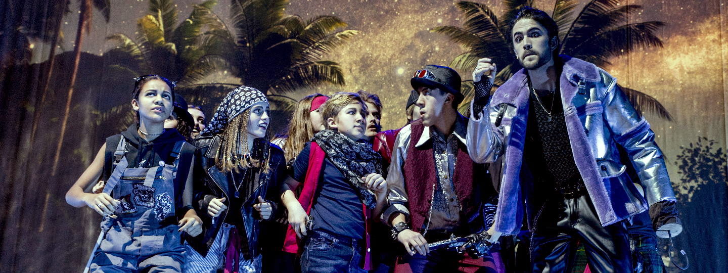 Eine Gruppe Jugendlicher steht auf einer Bühne, im Hintergrund zeigen Kulissen Palmen und einen Sternenhimmel. Die Jugendlichen scharen sich erwartungsvoll um einen größeren Mann mit Hakenhand, der Captain Hook verkörpert.