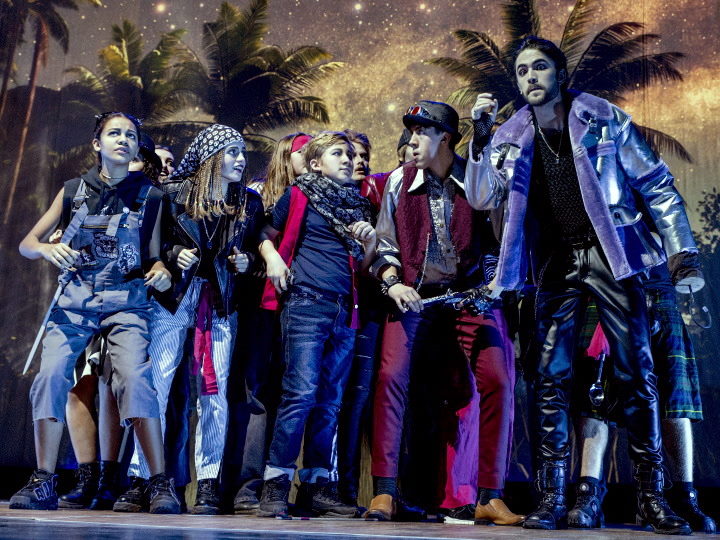 Eine Gruppe Jugendlicher steht auf einer Bühne, im Hintergrund zeigen Kulissen Palmen und einen Sternenhimmel. Die Jugendlichen scharen sich erwartungsvoll um einen größeren Mann mit Hakenhand, der Captain Hook verkörpert.
