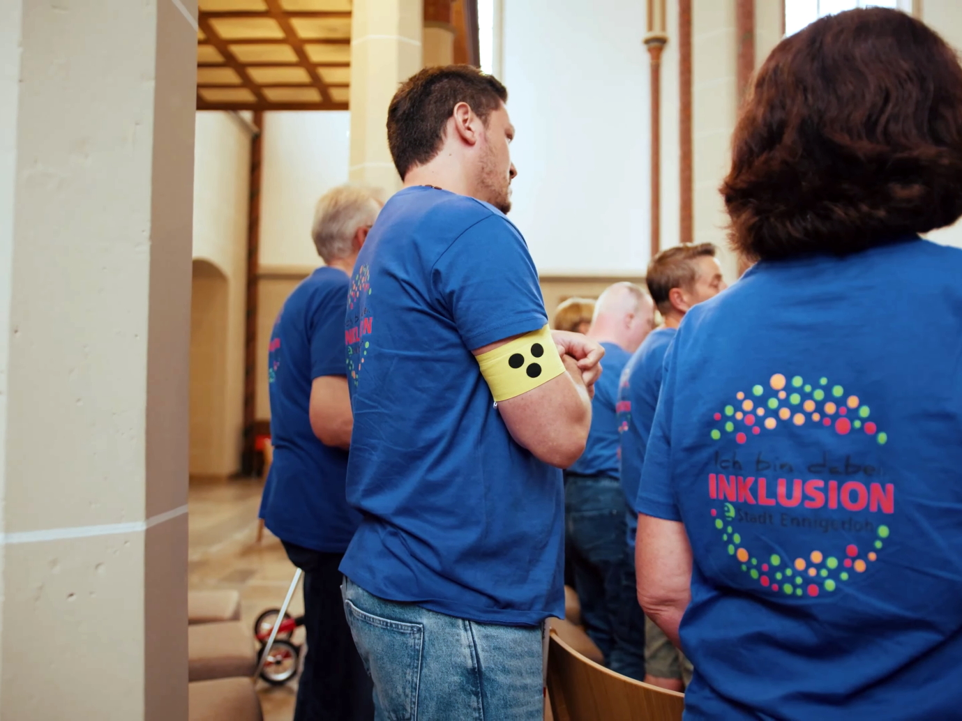 Neben einer Kirchensäule steht als Teil einer Gruppe, die in blaue T-Shirts gekleidet ist, ein Mann mit einer gelben Armbinde mit drei Punkten, wie sie von Menschen mit Sehbeeinträchtigung getragen wird.