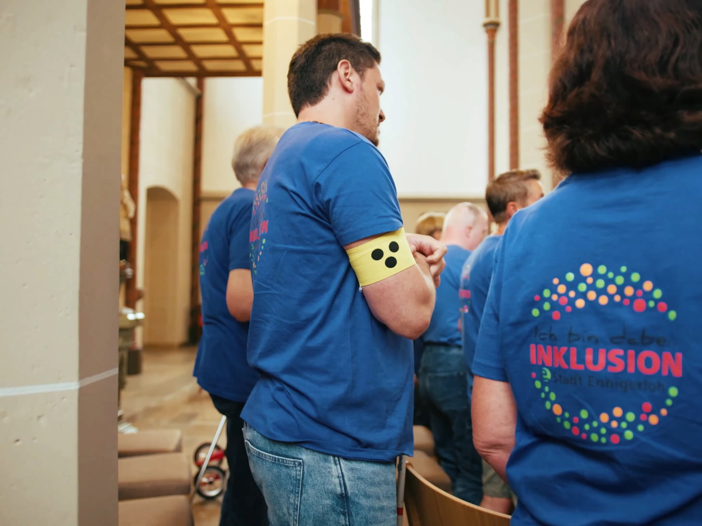 Neben einer Kirchensäule steht als Teil einer Gruppe, die in blaue T-Shirts gekleidet ist, ein Mann mit einer gelben Armbinde mit drei Punkten, wie sie von Menschen mit Sehbeeinträchtigung getragen wird.