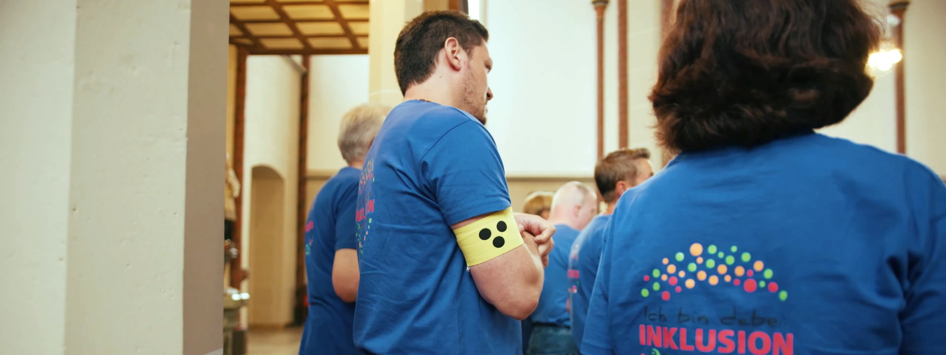 Neben einer Kirchensäule steht als Teil einer Gruppe, die in blaue T-Shirts gekleidet ist, ein Mann mit einer gelben Armbinde mit drei Punkten, wie sie von Menschen mit Sehbeeinträchtigung getragen wird.