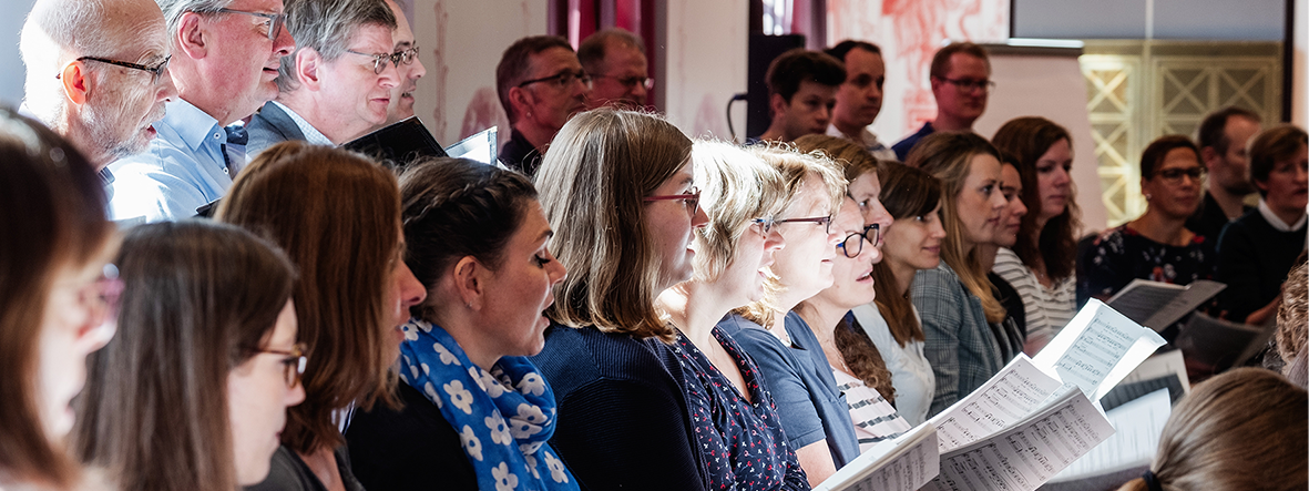 Etwa 10 Männer und Frauen singen zusammen. Einige halten Noten in den Händen. Die Blicke gehen alle in dieselbe Richtung, zur dirigierenden Person