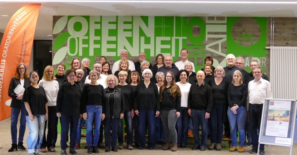 36 Menschen unterschiedlichen Alters stehen vor einer grünen Wand, auf der Offen und Offenheit steht. Vorne im Bild steht ein Poste mit der Aufschrift Traum und Sehnsucht