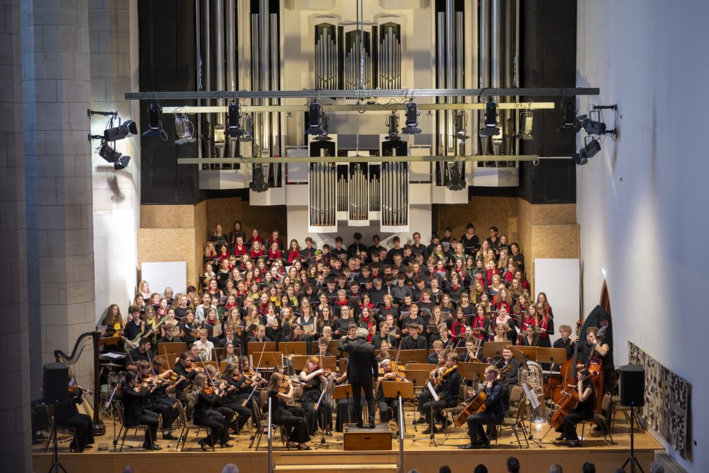 Etwa 80 Menschen stehen auf einer Bühne und singen. Sie tragen schwarze Kleidung mit roten, grünen und gelben Schals. Im Vordergrund sitzen etwa 20 Musikerinnen und Musiker, die im Orchester Instrumente spielen. Davor steht ein Dirigent. Im Hintergrund ist eine große Orgel mit vielen Orgelpfeifen zu sehen.