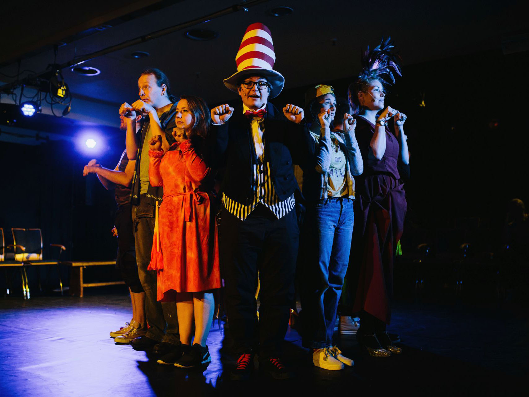 In der MItte einer Bühne steht eine Person mit rot-weiß geringeltem Zylinder auf dem Kopf. Links und rechts stehen 4 weitere Menschen und blicken suchend in die Gegend