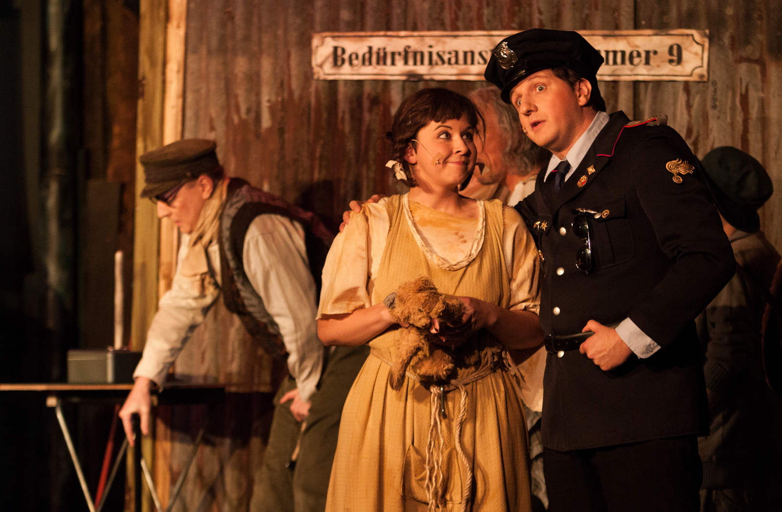 Zwei Schauspieler im Fokus: Ein Polizist und ein kleines Mädchen stehen vor einer Bedürfnisanstalt aus dem Musical Pinkelstadt. Er schaut erstaunt, sie lächelt.