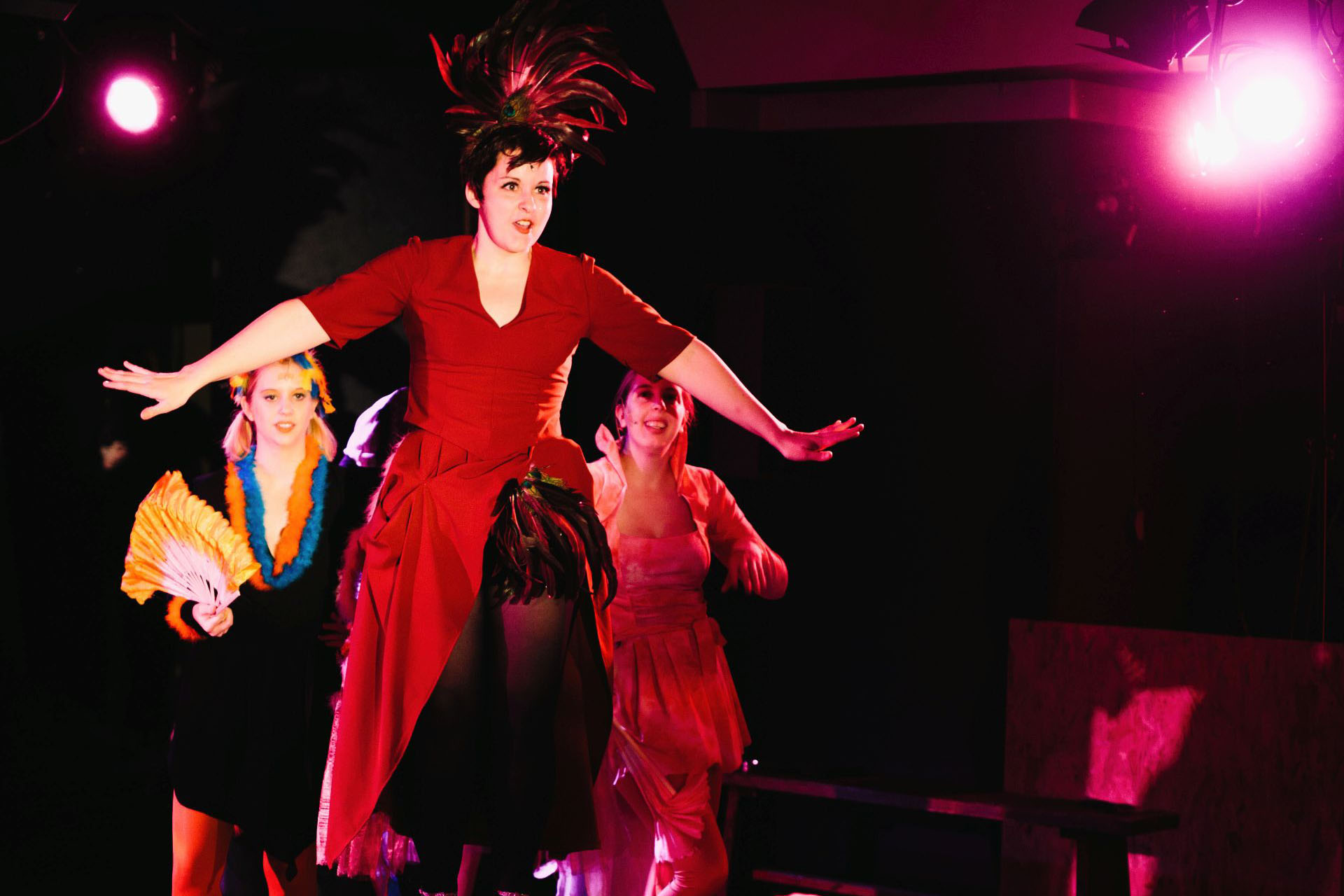 Eine Frau mit rotem Kleid und Federn auf dem Kopf steht vor zwei weiteren Menschen. Ihre Arme sind ausgebreitet. Sie blickt entschlossen nach vorne.