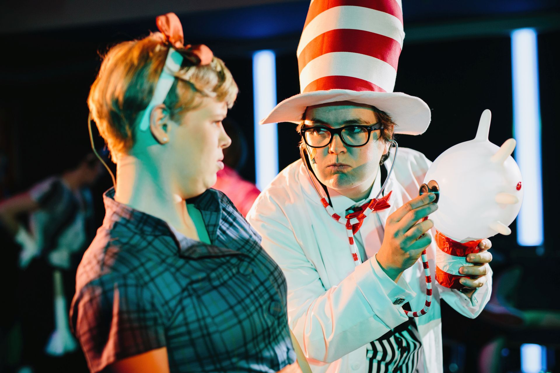 Eine Person in weißem Kittel und rot-weiß geringeltem Zylinder hält ein Stethoskop an einen Luftballon. Eine zweite Person schaut skeptisch auf das Geschehen.
