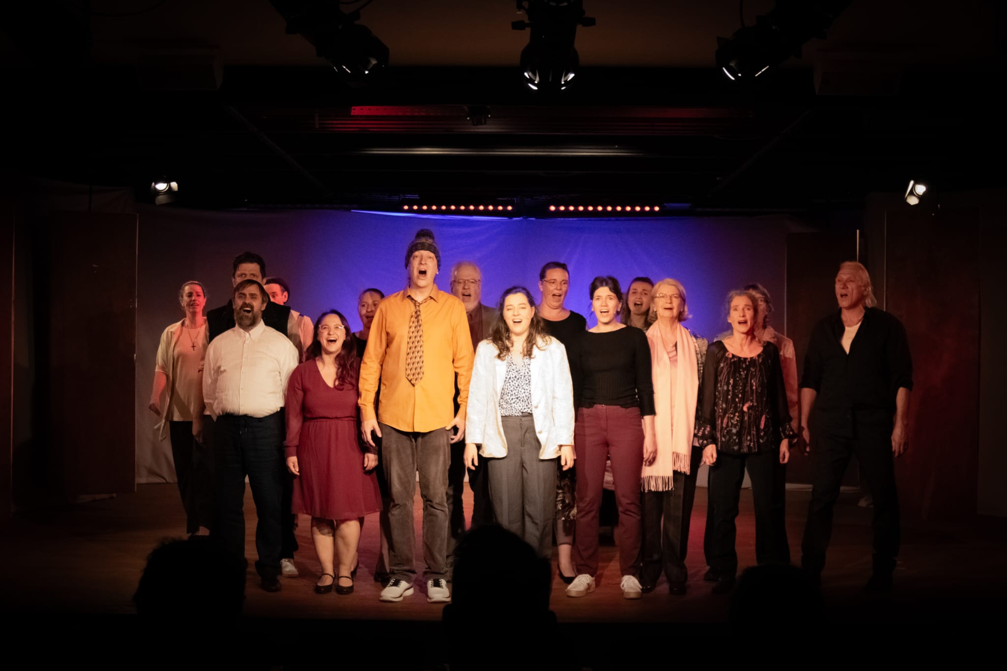 Etwa 15 Menschen stehen in Alltagskleidung auf der Bühne und singen