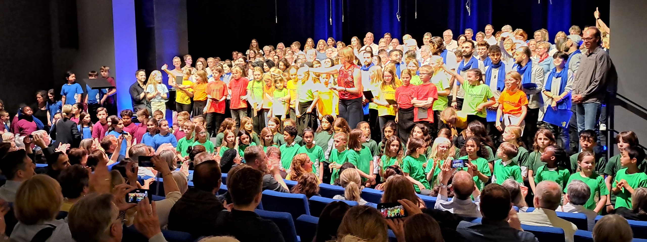 eine Bühne voller Kinder in bunten T-Shirts und begeisterte Menschen im Publikum, die ihre Handys zücken um Fotos zu machen