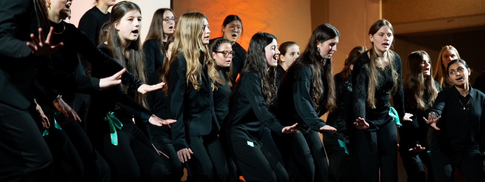 Etwa zwanzig Mädchen aus dem Mädchenchor der Singakademie zu Berlin singen auf einer Bühne vor orange und weiß angestrahlten Wänden. Sie tragen schwarze Konzertkleidung und unterstützen mit ausgestreckten Händen ihren Gesang.