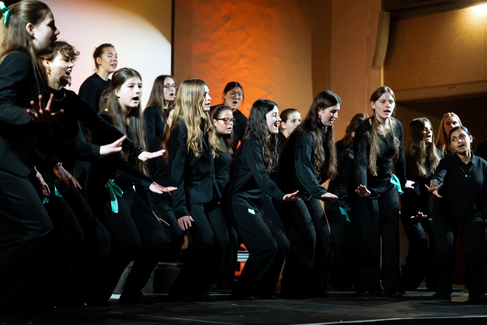 Etwa zwanzig Mädchen aus dem Mädchenchor der Singakademie zu Berlin singen auf einer Bühne vor orange und weiß angestrahlten Wänden. Sie tragen schwarze Konzertkleidung und unterstützen mit ausgestreckten Händen ihren Gesang.