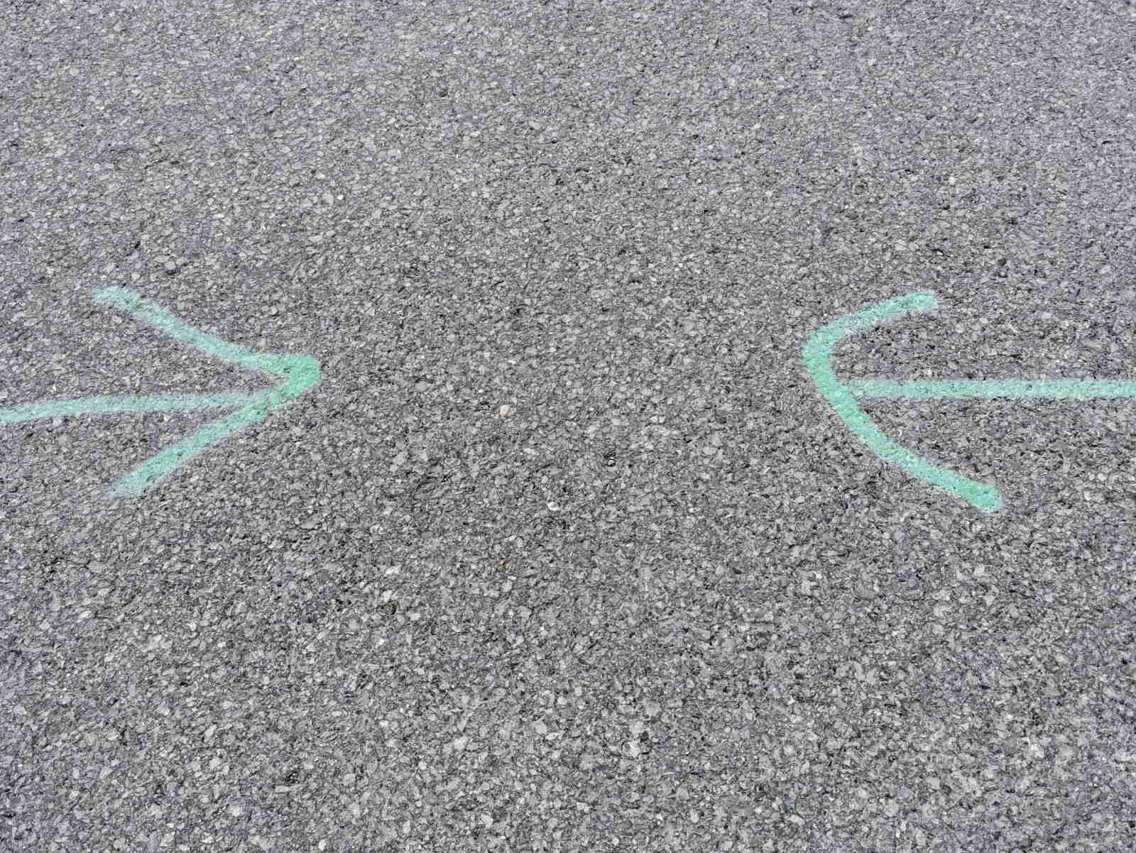 Auf einer grauen Asphaltfläche sind mit grüner Kreide zwei Pfeile gezeichnet, die aufeinander zeigen.