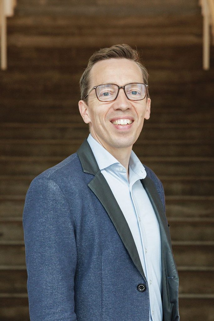 Stephan Doormann lächelt, leicht seitlich eingedreht, der betrachtenden Person entgegen. Er hat kurze braune Haare, eine rechteckige Brille und trägt dunkelblaues Sakko über hellblauem Hemd.