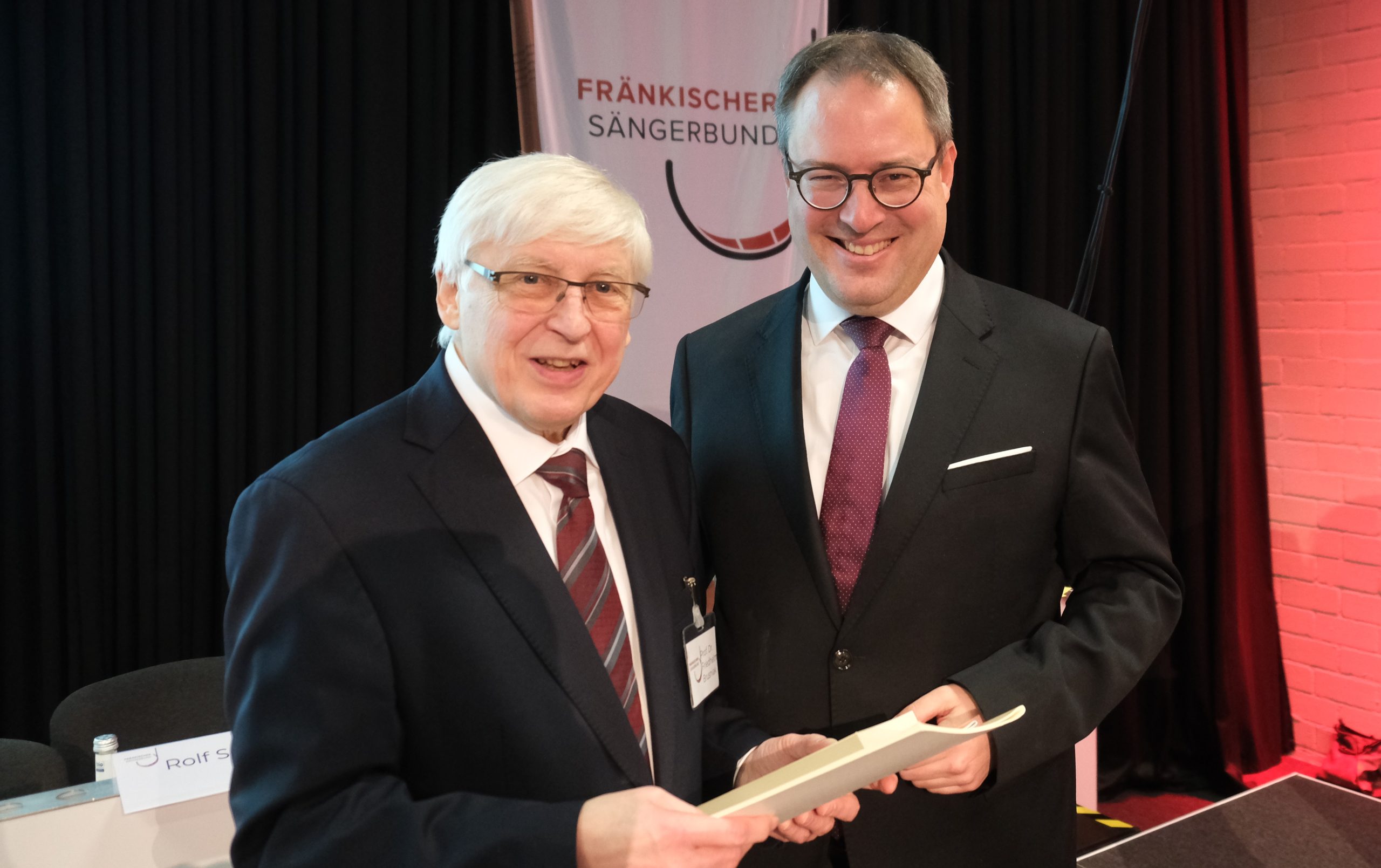 Ein Mann mit weißen Haaren steht neben einem Mann mit dunklen kurzen Haaren. Beide lächeln in die Kamera