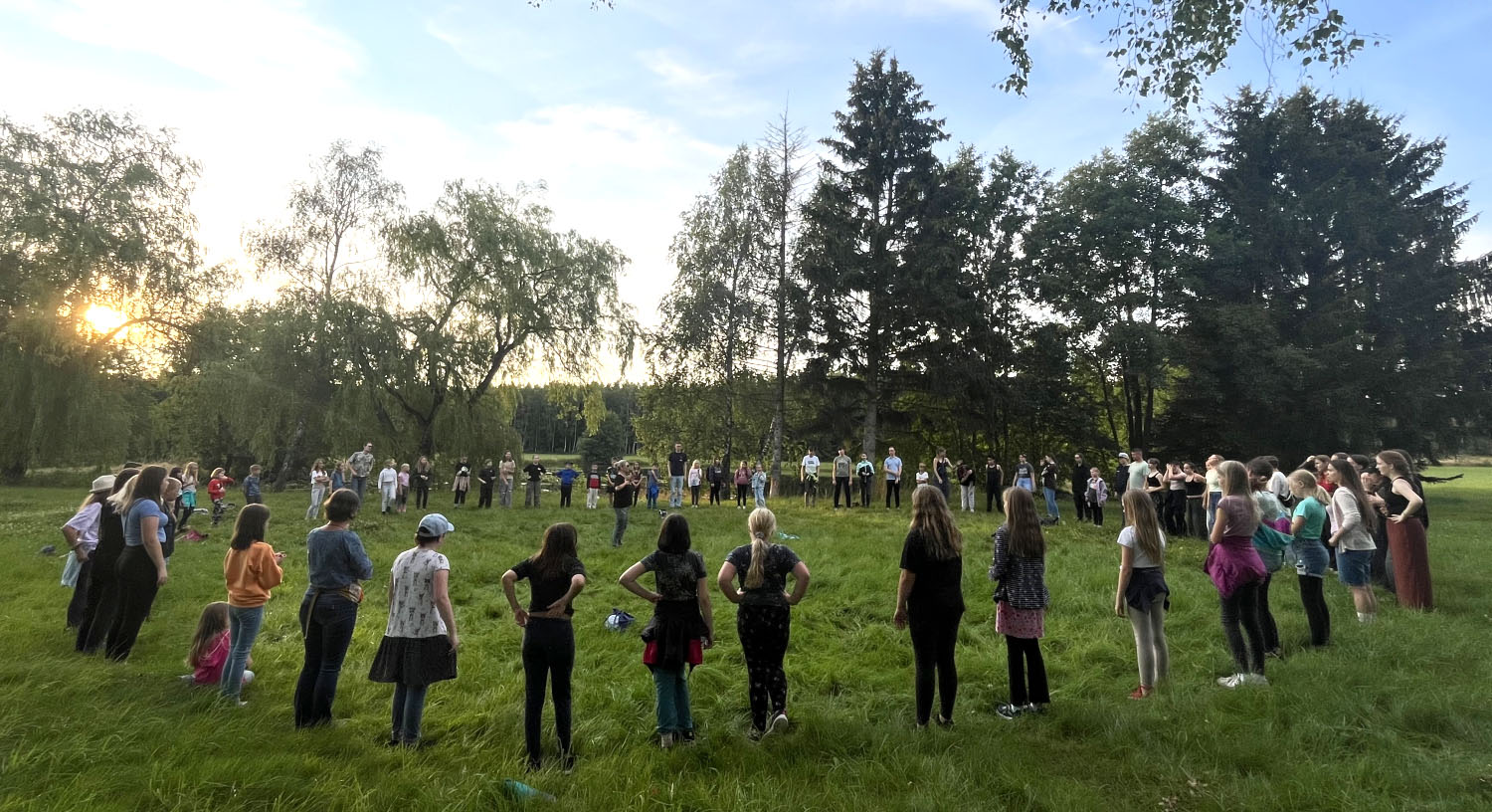 Etwa 50 Kinder und Jugendliche stehen auf einer Wiese im Kreis. In der Mitte liegt ein Ball. Im Hintergrund scheint die Sonne durch die Bäume