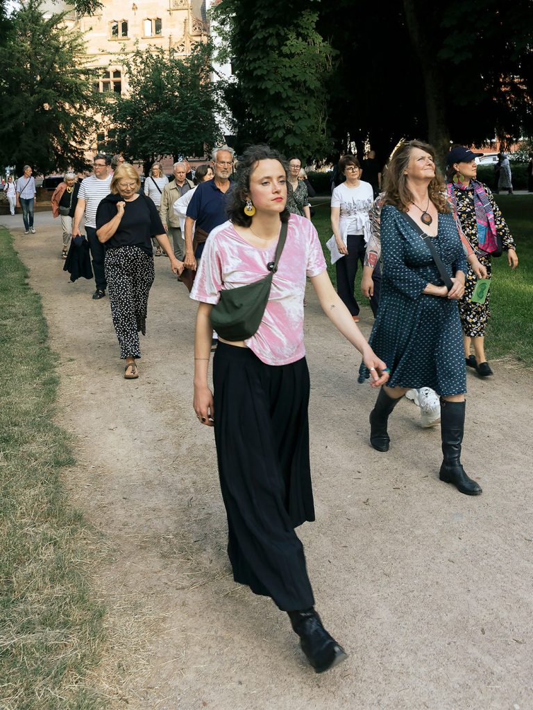 Die Kuratorin Sophie Emilie Beha läuft an der Spitze eines kleinen Menschenzugs auf einem Kiesweg durch den Kölner Stadtgarten.